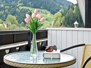 Een ronde glazen tafel met een vaas roze bloemen, een boek en zonnebril, met uitzicht op een schilderachtig bewoording van een bebost dal.