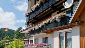 En traditionell chalet med träbalkonger prydda med blommor, en vit fasad och en bergskuliss i bakgrunden.
