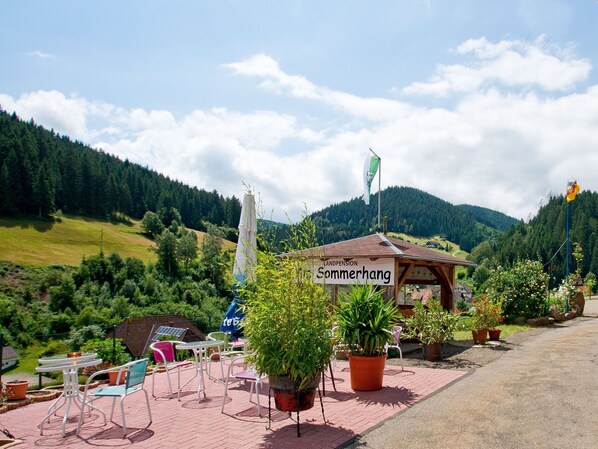 Un petit espace extérieur aménagé avec des tables et des chaises, entouré de plantes en pot, sous une enseigne où l’on peut lire « Sommerhang ».
