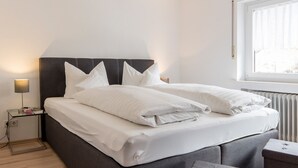 A neatly made bed with white linens and pillows, a bedside table with a lamp, and a window with blinds. - Apartment 'Rosi Mit 80qm' with Shared Terrace, Balcony and Wi-Fi (Bad Rippoldsau-Schapbach)