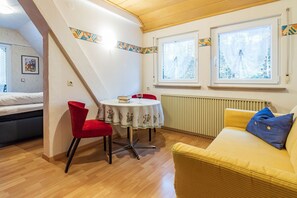 Um quarto com cama, uma cadeira vermelha, uma mesa com toalha e um sofá amarelo, além de duas janelas com cortinas.