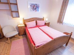 A bedroom with a wooden bed, a nightstand, a chair, a red rug, a window with curtains, and a painting on the wall.