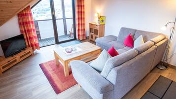 A living room with a sofa, a coffee table, a TV on a stand, a small wooden cabinet, and a view through a large window.