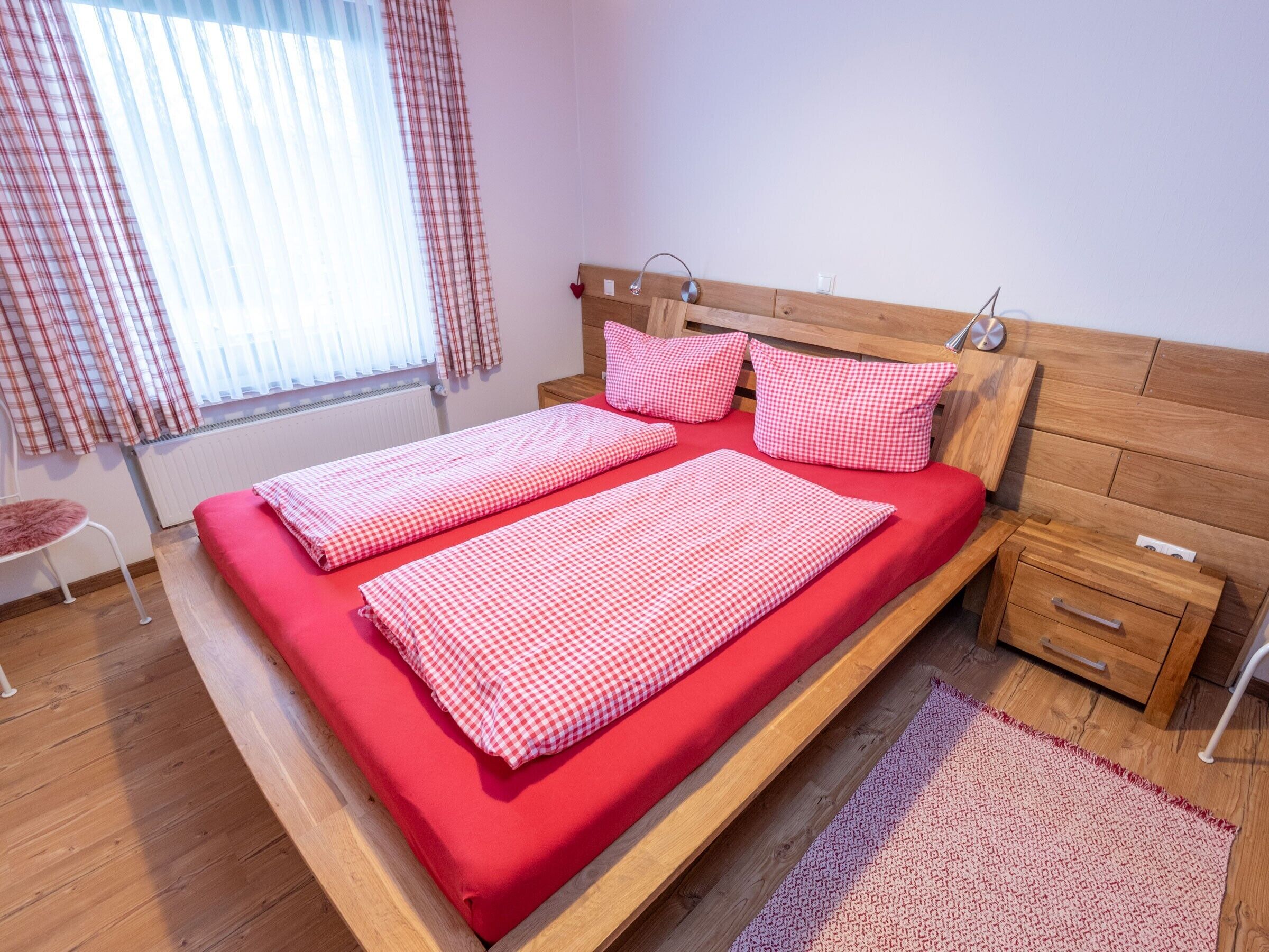 A double bed with red bedding, a wooden headboard, a bedside table with a drawer, and a window with curtains.