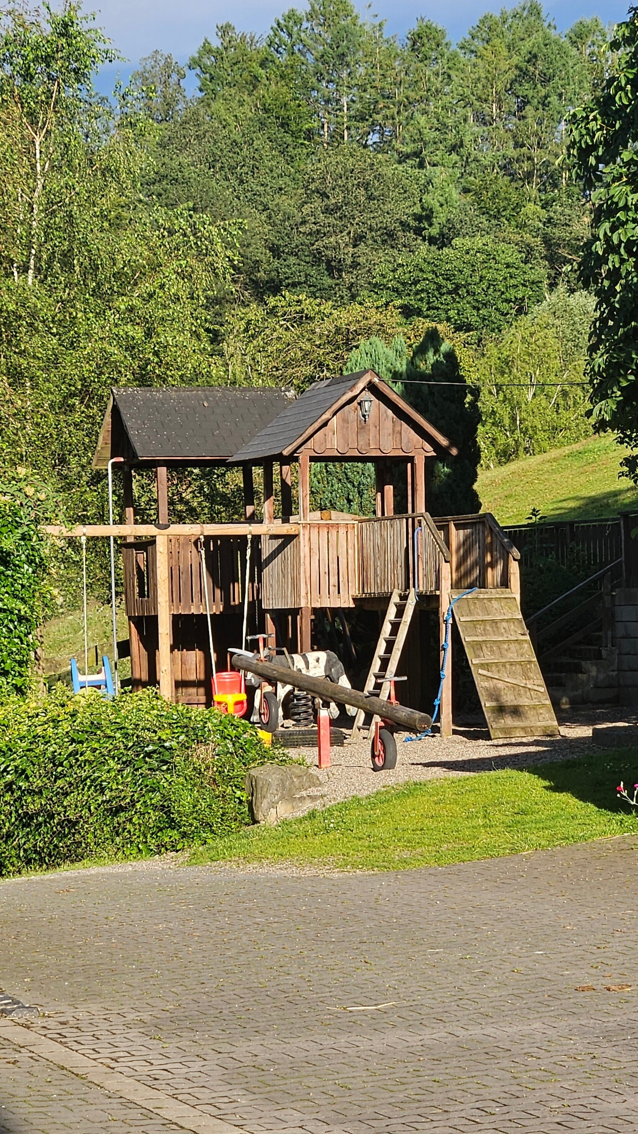 Ein Spielhäuschen aus Holz mit einer Rampe, einer Schaukel und einer Rutsche.