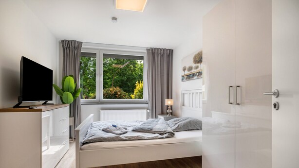 Un dormitorio con una cama, un televisor, una ventana con cortinas, una planta y un armario.