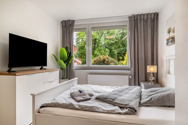 A bedroom with a bed, a TV on a wooden cabinet, a window with curtains, a bedside lamp, and a potted plant.