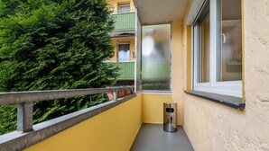 Un balcón con barandilla metálica, un cubo de basura pequeño y una ventana con vistas a un follaje verde y frondoso.