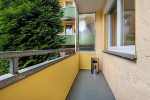 Uma varanda com guarda-corpo de metal, uma pequena lixeira e uma janela com vista para folhagem verde densa.