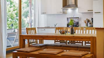 Una mesa de comedor de madera con bancos acolchados, un jarrón con flores y vista a una cocina con armarios blancos y una ventana.