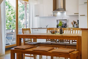 Une table à manger en bois avec des bancs rembourrés, un vase rempli de fleurs et une vue sur une cuisine avec des meubles blancs et une fenêtre.