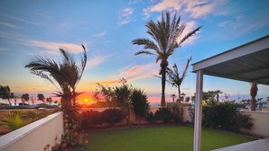 A patio area with a view of the sunset, palm trees, and a grassy lawn. - Gugel Waves Seaview 4BR Family Aparts (Nahariya)