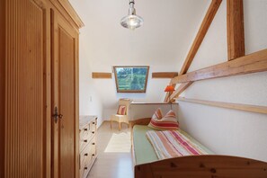 Quarto com cama, guarda-roupa de madeira, uma cadeira, escrivaninha com abajur e janela com vista para área verde.