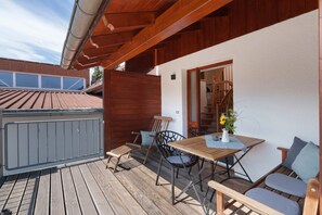 Um deck de madeira com mesa e cadeiras, uma mesinha com um vaso de flores e vista para um edifício com telhado vermelho.