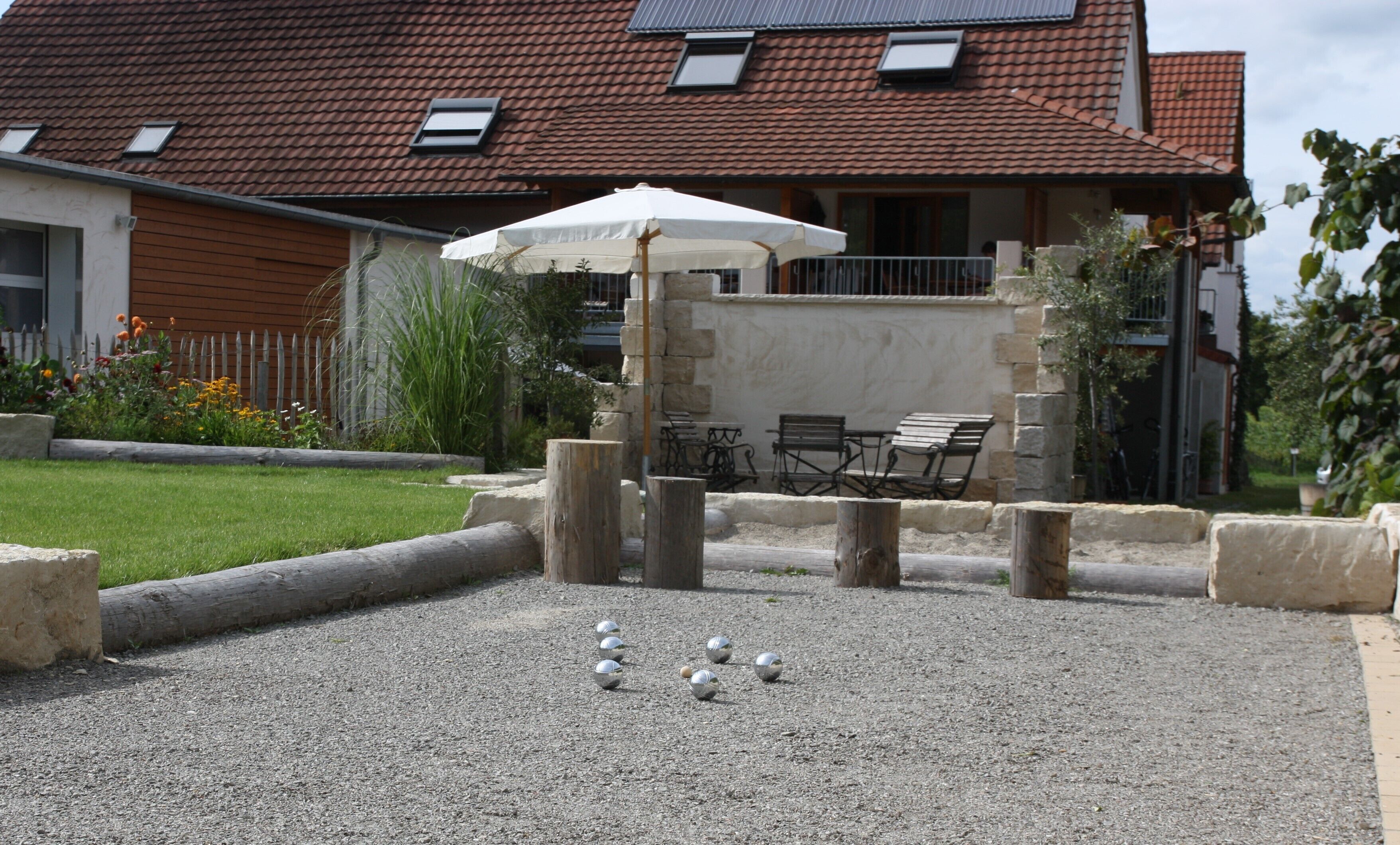 Eine Terrasse mit Tisch und Stühlen, einem weißen Sonnenschirm und einem Kiesbelag mit Metallkugeln.