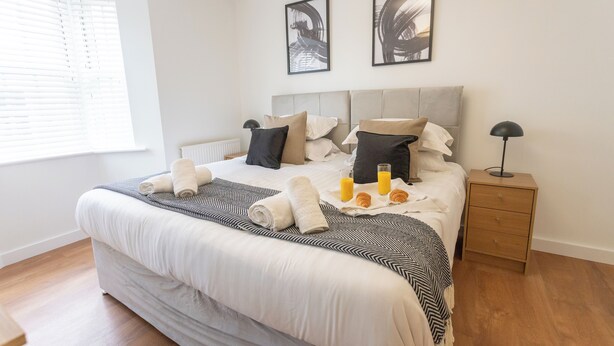 Ein ordentlich bezogenes Bett mit einer gestreiften Decke, Kissen und zusammengerollten Handtüchern. Zwei Nachttische, einer mit einer Lampe und der andere mit einer Schublade. Ein Fenster mit Jalousien ist sichtbar, und zwei gerahmte Bilder hängen an der Wand.