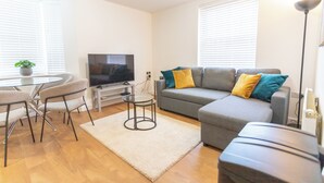 Ein modernes Wohnzimmer mit grauem Sofa, einem Glastisch, einem Fernseher und einem kleinen Beistelltisch.