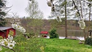 Una propiedad tranquila junto al lago con una cabaña roja, abundante vegetación y flores blancas en flor.