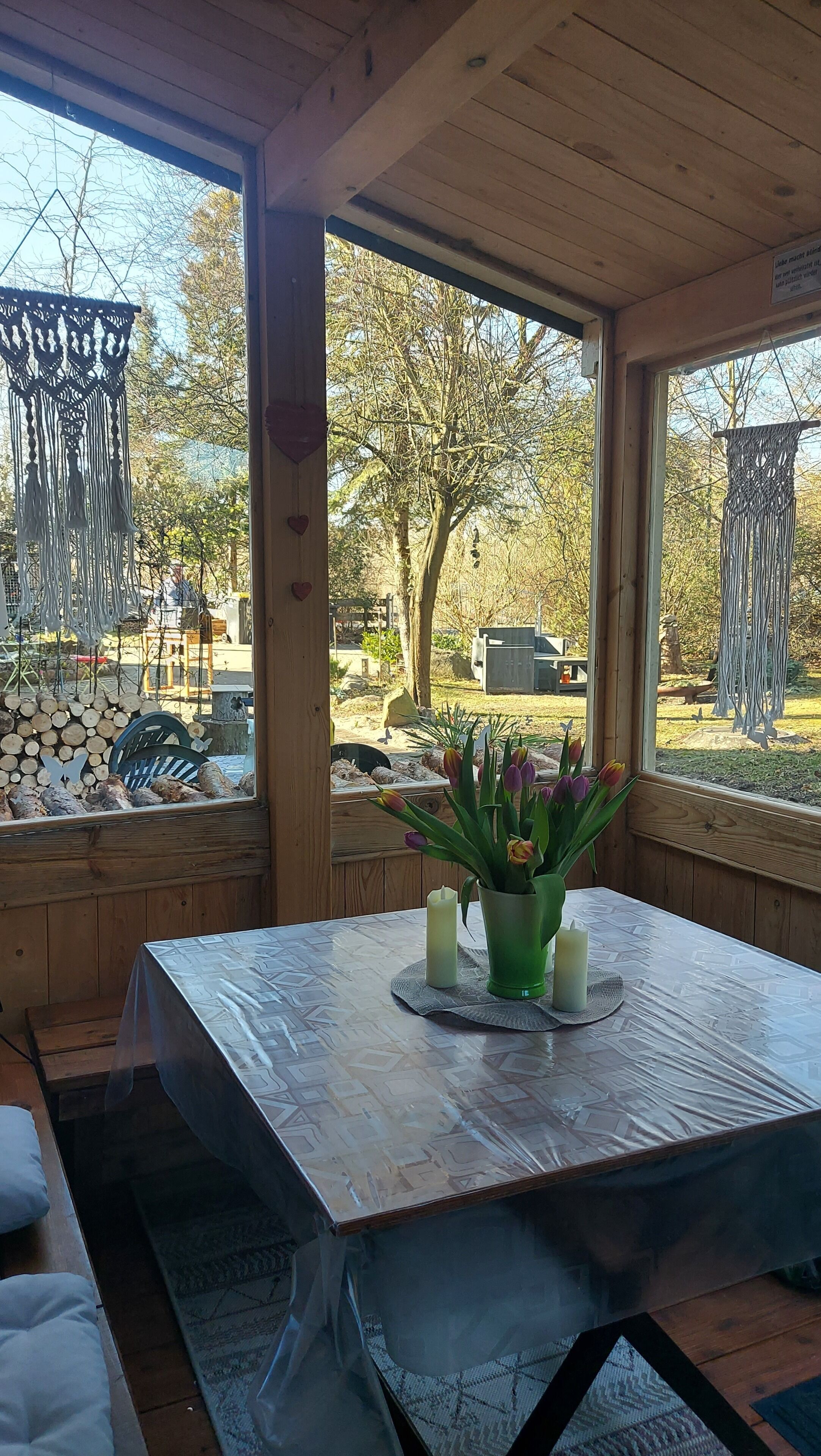 A wooden interior with a table covered by a plastic tablecloth, a green vase with flowers, and candles.