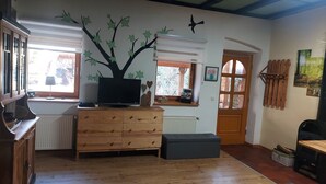 Una habitación con un aparador de madera, un televisor encima, un radiador, una ventana con persianas, una puerta y una pared con un adhesivo de árbol.