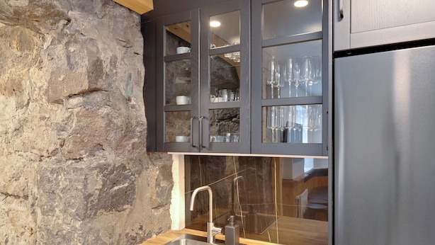 Une cuisine avec un mur en pierre, un évier en acier inoxydable et des meubles de rangement sombres dotés de portes en verre.
