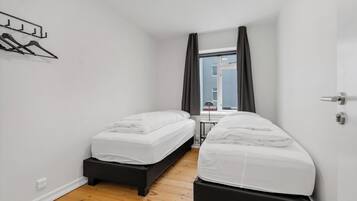 Um quarto pequeno e moderno com duas camas de solteiro, piso de madeira, janela com cortinas e um cabideiro fixado na parede.