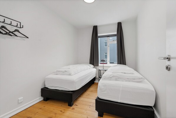 A small, modern bedroom with two single beds, a wooden floor, a window with curtains, and a wall-mounted coat rack. - Ranargata Apartments by Heimaleiga (Reykjavík)