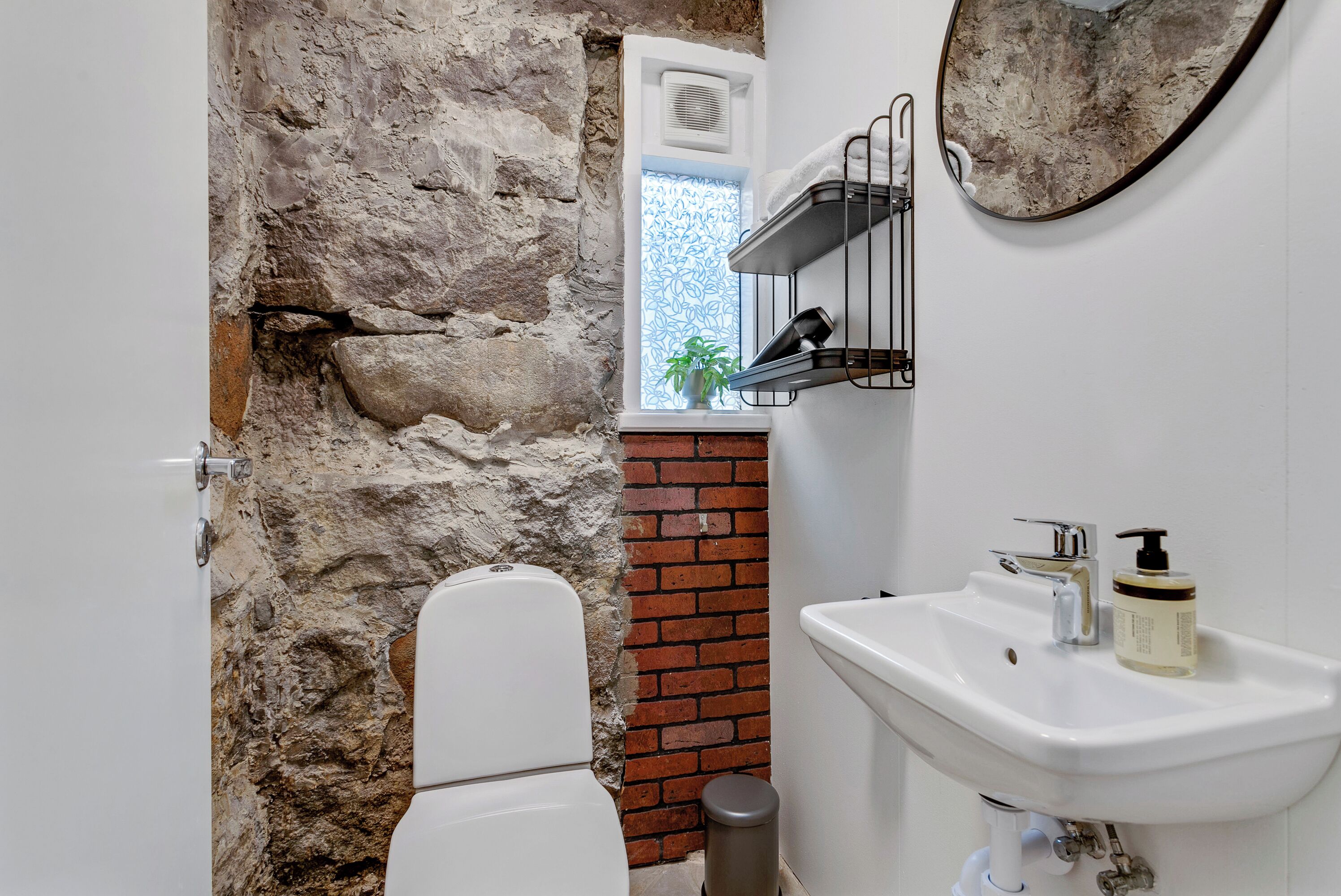 A bathroom with a stone wall, a white toilet, a sink with a mirror, and a window with a plant.