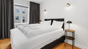 Un lit bien fait, avec du linge de lit blanc, une tête de lit sombre et une table de chevet sur laquelle se trouve une lampe.