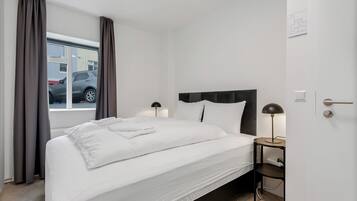 A neatly made bed with white linens, a bedside table with a lamp, and a window with curtains.
