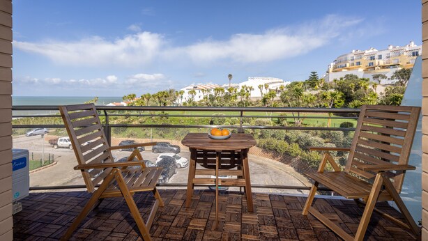 Ein Balkon mit zwei Holzstühlen und einem Tisch, einer Obstschale und Blick auf eine Küstenlandschaft mit Gebäuden und Grünflächen.