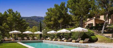 A swimming pool with lounge chairs and umbrellas, surrounded by trees and a mountain in the background.