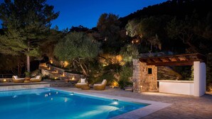A swimming pool with lounge chairs and a stone structure, surrounded by trees and a hillside.
