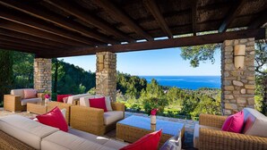 An outdoor seating area with wicker furniture and stone pillars, overlooking a scenic view of the sea and hills.