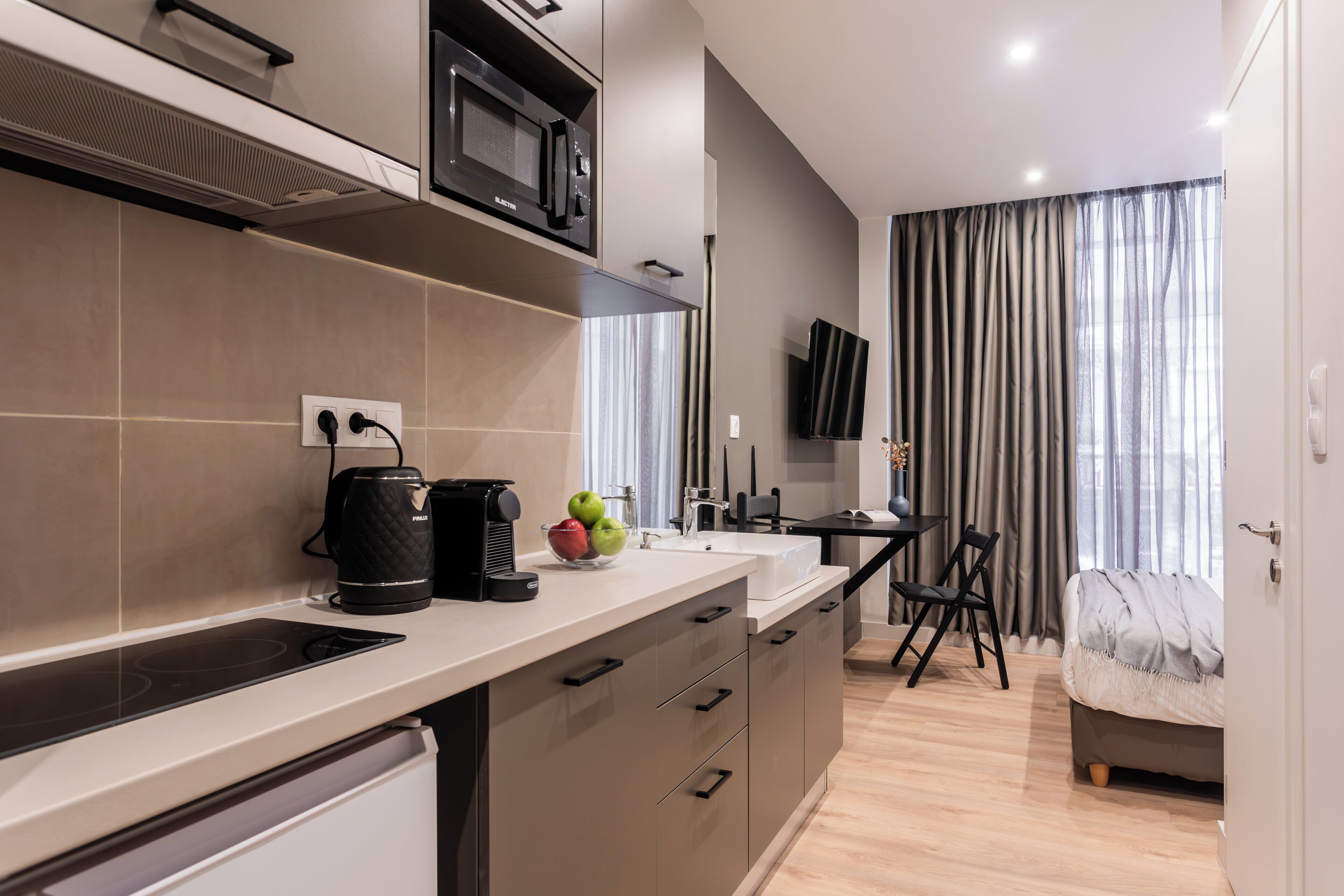 A modern kitchen with a built-in microwave, a coffee maker, and a fruit bowl. There is a dining area with a table and chairs, and a TV mounted on the wall.