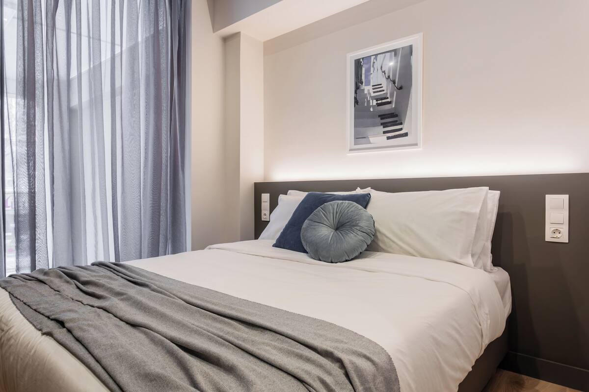 A neatly made bed with a grey blanket, white pillows, and a blue accent pillow. A framed picture hangs on the wall above the bed.