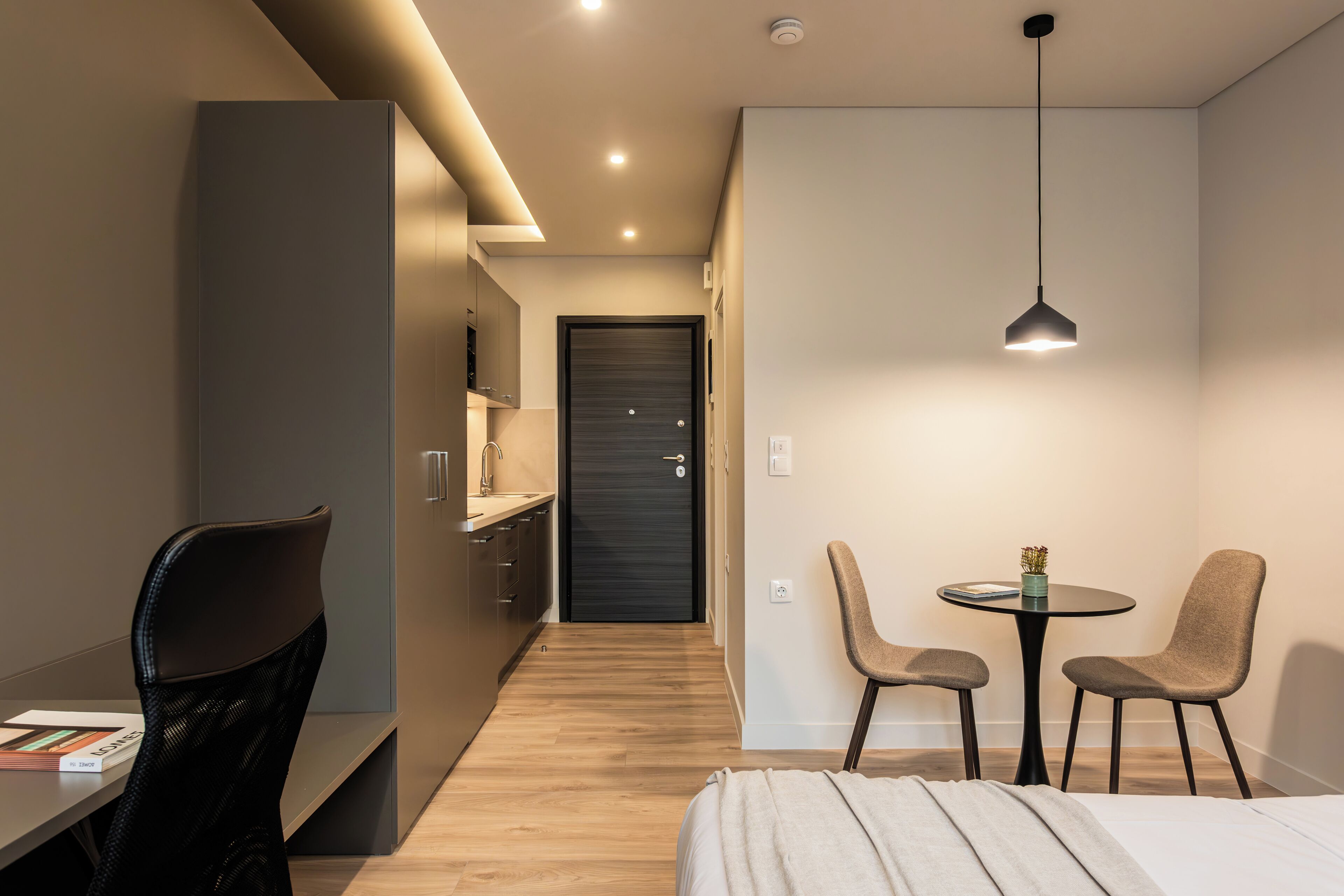 A modern hotel room with a bed, a desk, a chair, a small table with two chairs, and a door leading to the bathroom.