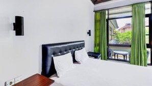 A hotel room with a bed, a nightstand, a wall-mounted lamp, and a view of the outdoors through a window with green curtains. - Anna Kuta Inn Hotel (Kuta)