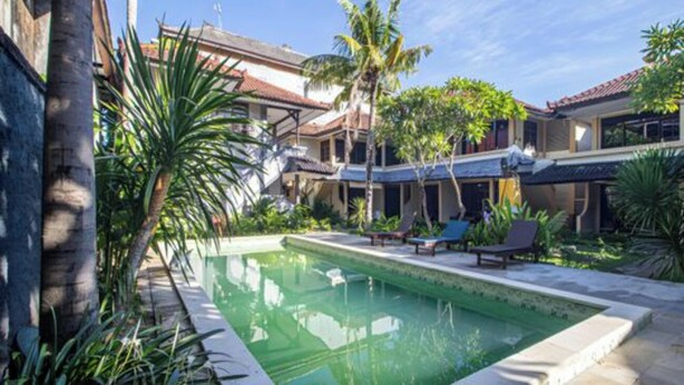 A swimming pool with lounge chairs, surrounded by palm trees and a building with a tiled roof. - Anna Kuta Inn Hotel (Kuta)