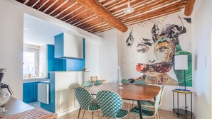 A dining area with a wooden table and patterned chairs, a kitchen with blue cabinets, and a mural on the wall.