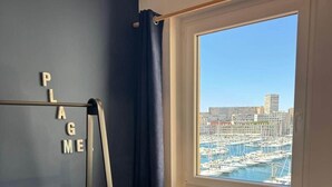 A room with a view of a marina, a desk, a chair, a window with curtains, and a metal rack with letters spelling 'PLAGUE'.