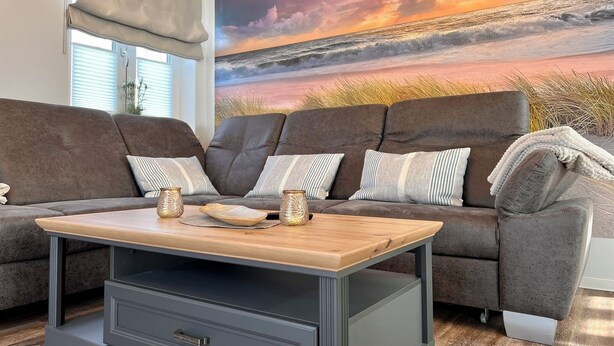 Un salon avec un canapé gris, deux coussins, une table basse en bois et un grand tableau représentant un coucher de soleil sur l’océan.