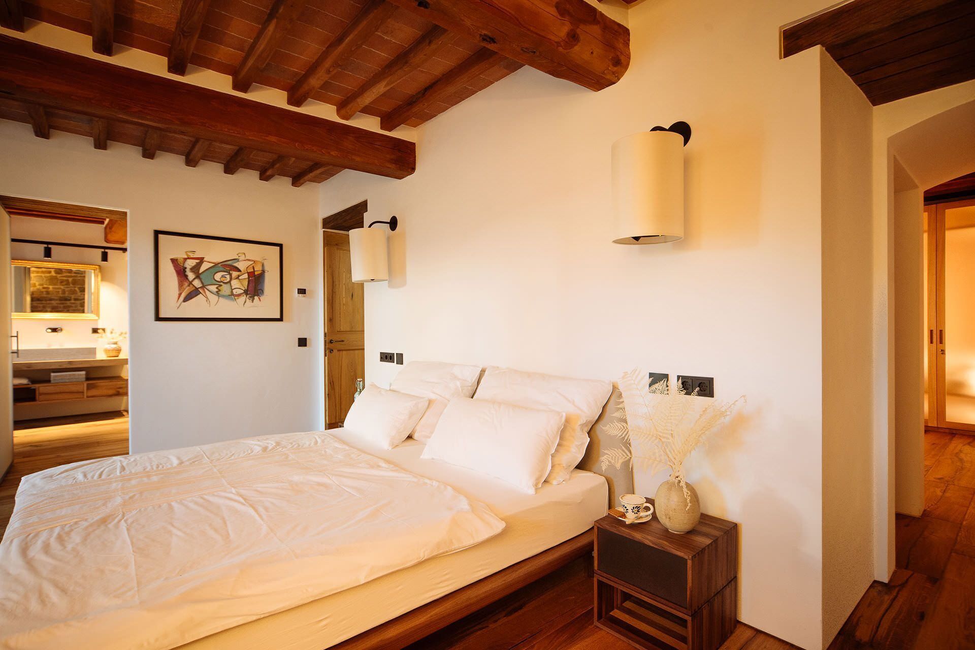 A bedroom with a wooden bed, white bedding, a bedside table with a vase, and a wall-mounted lamp.