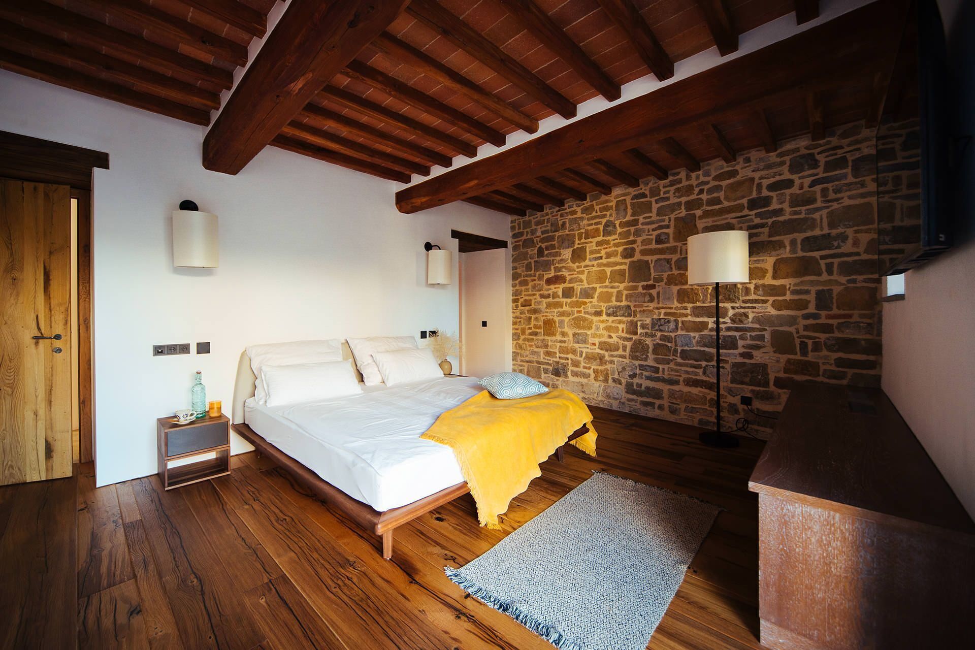 Une chambre avec un plafond en bois, un mur en pierre, un lit avec une literie blanche, une table de chevet, une lampe et un plancher en bois.