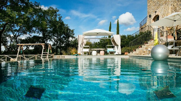 A clear blue pool with a wooden deck, lounge chairs, and a stone building in the background. - My Rental Homes - Villa Torre del Chianti with hited pool, Gym and wellness center (Radda In Chianti)