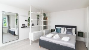 Un dormitori modern amb un llit gran, un armari blanc, un mirall i una estanteria amb plantes.