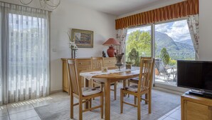 Un comedor con mesa, sillas, televisor y vistas a montañas a través de amplios ventanales.