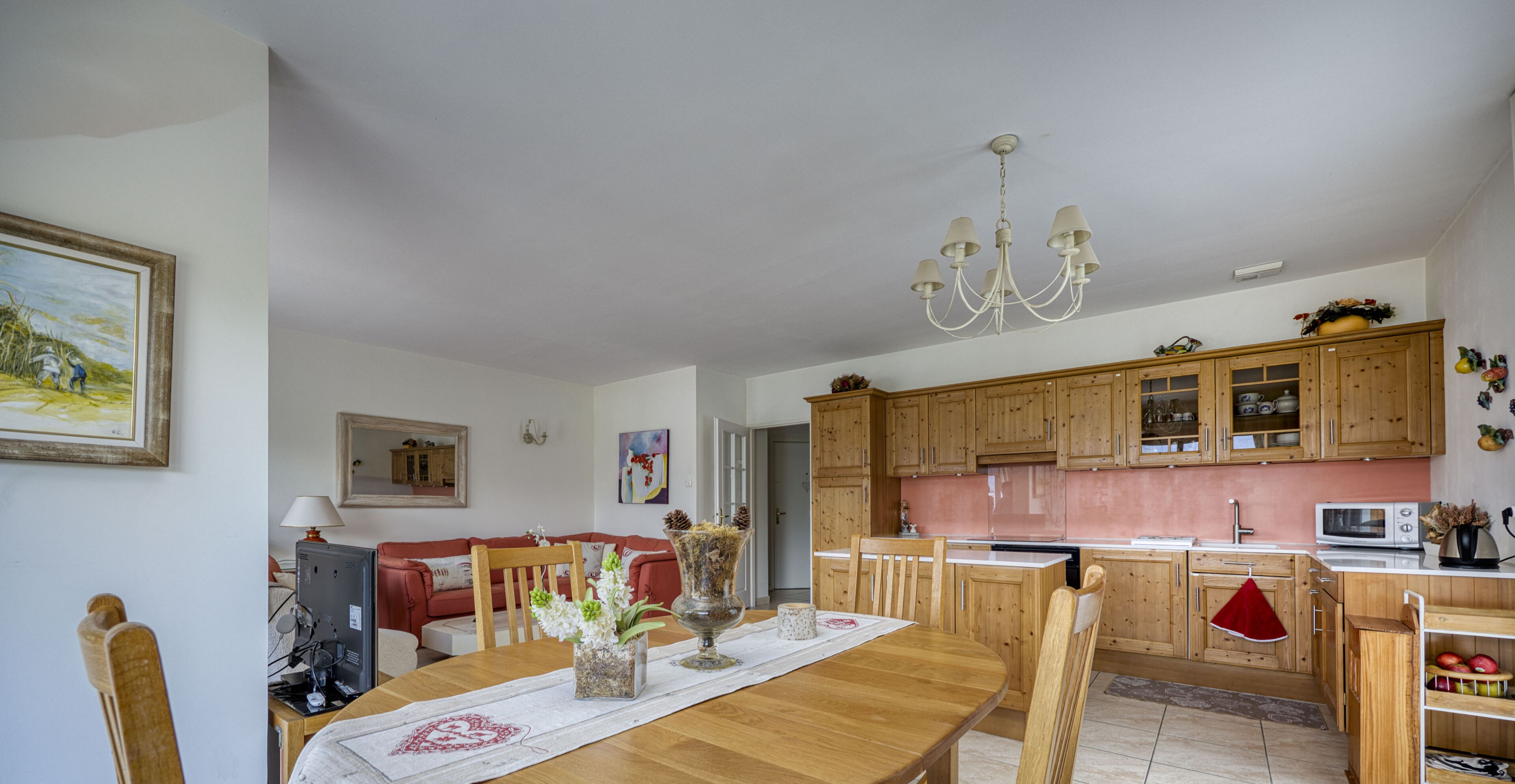 On aperçoit un coin repas avec une table en bois, des chaises et un lustre. Au fond, on distingue une cuisine avec des meubles en bois et un four à micro-ondes.