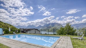 Una piscina con montañas al fondo, un edificio pequeño y un área cercada con un banco.