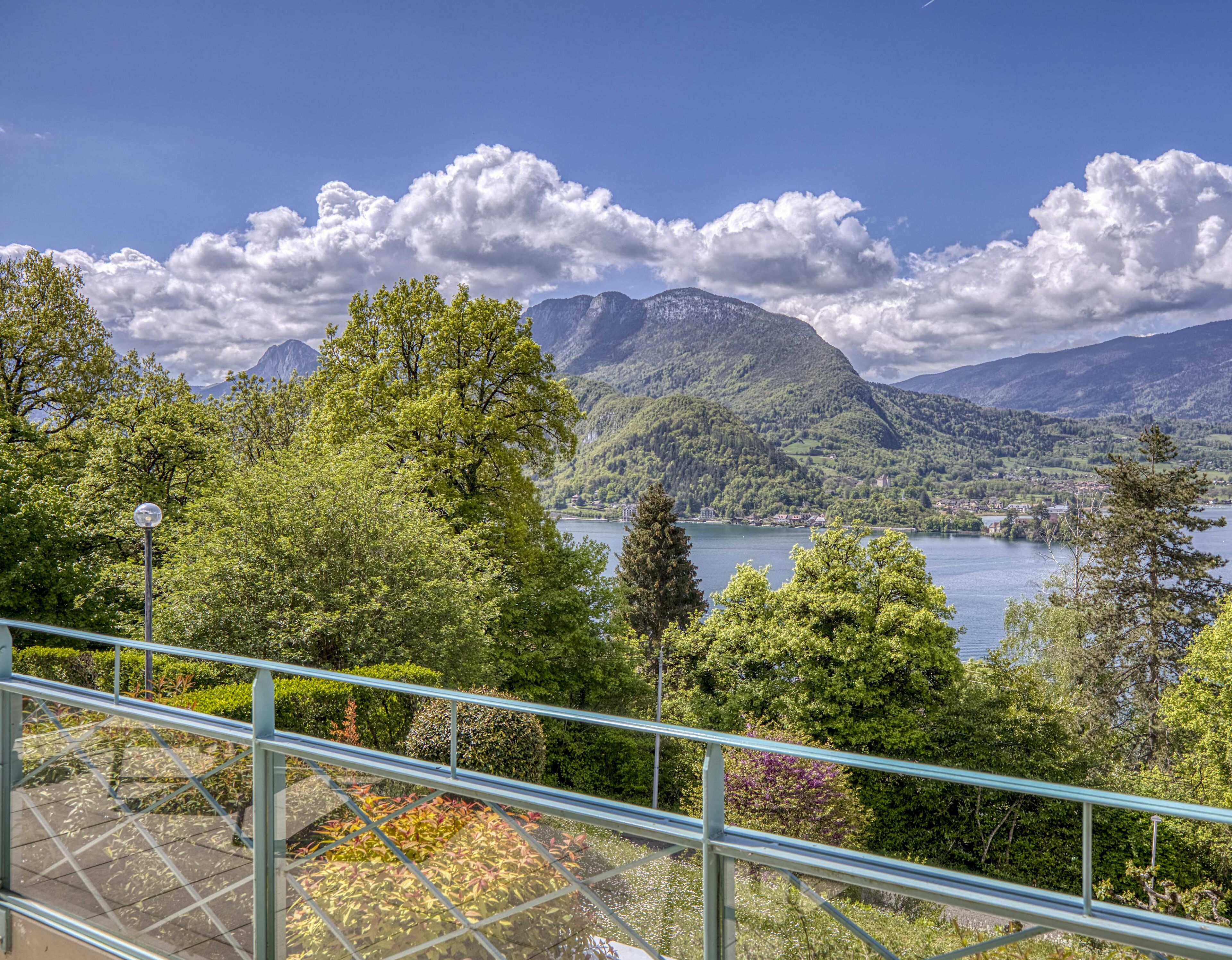 Ein malerischer Blick auf einen See mit Bergen im Hintergrund, einem Metallgeländer im Vordergrund und üppiger Vegetation.